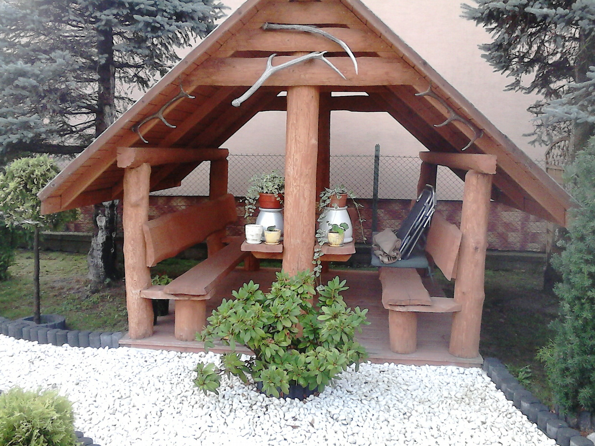 Drewniana altana ogrodowa z miejscami do siedzenia i stołem, ozdobiona porożem na dachu, otoczona białymi kamieniami i zielenią.