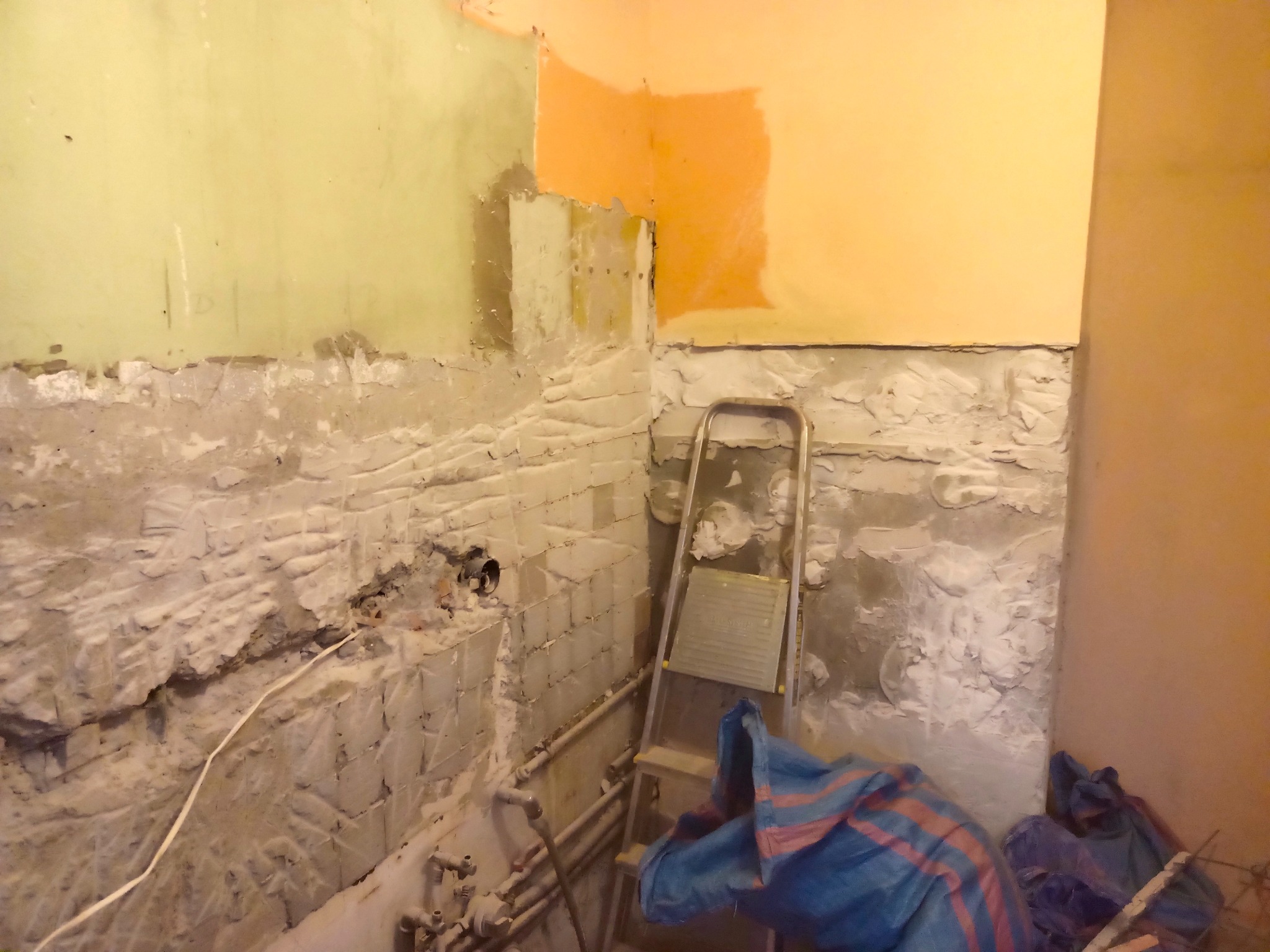 Ściana w trakcie przygotowania do prac wykończeniowych, widoczne fragmenty starej farby, odsłonięta cegła, rury instalacyjne i drabina aluminiowa z plandeką.