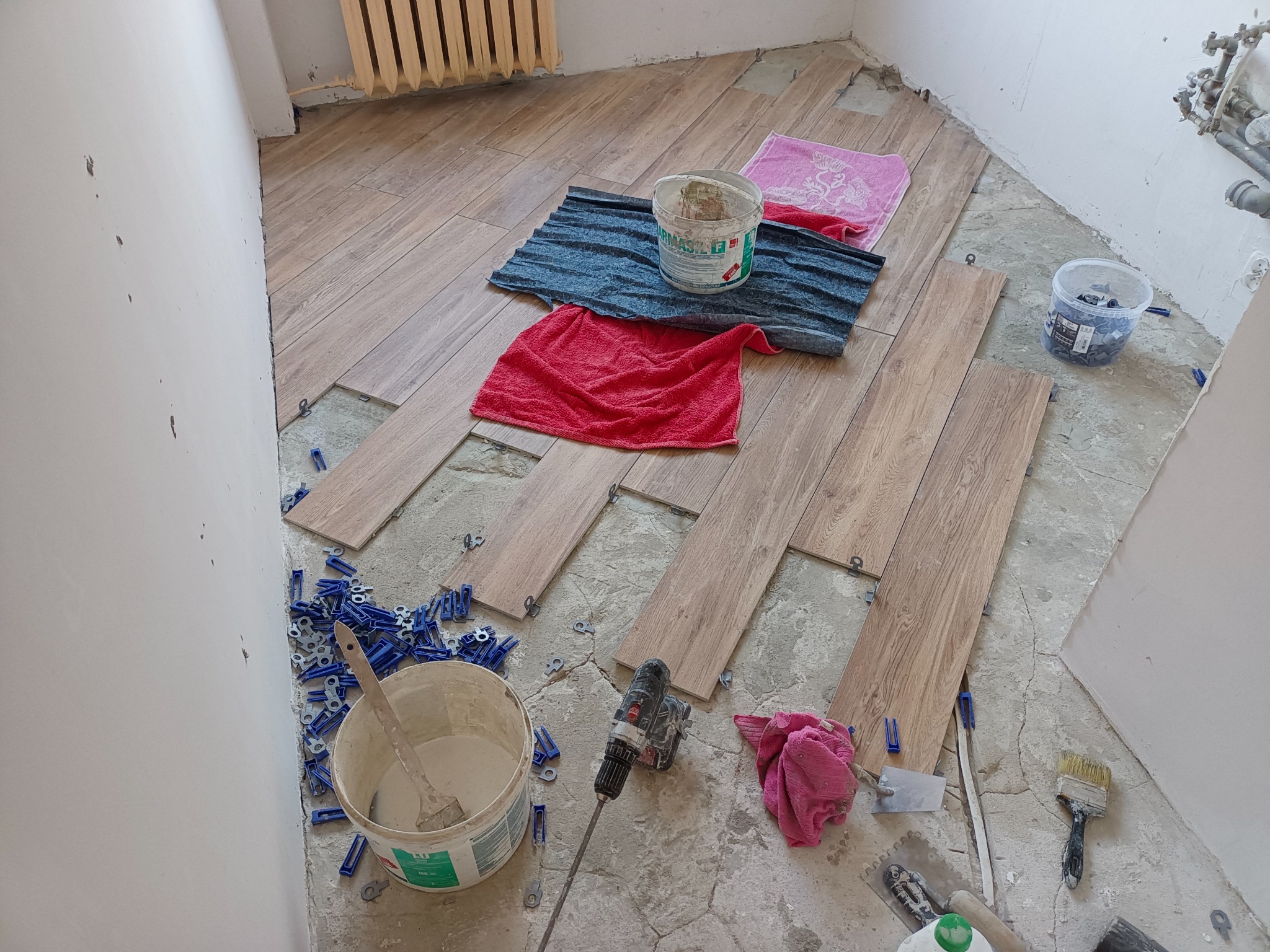 Układanie drewnopodobnych płytek podłogowych w pomieszczeniu, widoczne narzędzia glazurnicze, wiadro z klejem, poziomica, wiertarka z mieszadłem i kolorowe ścierki na podłodze.