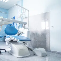 Niebieski fotel dentystyczny z oprzyrządowaniem w jasnym gabinecie, widok na ścianę z szarą mozaiką i gniazdkami elektrycznymi.