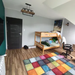 Pokój dziecięcy z drewnianym łóżkiem piętrowym, kolorowym dywanem w kwadraty, biurkiem z komputerem i zieloną ścianą z telewizorem.