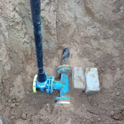 Niebieski zawór z żółtymi elementami, połączony z czarną rurą, zainstalowany w wykopie w Grzebienisku, zabezpieczony dwoma betonowymi bloczkami.