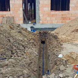 Wykopany wąski rów z ułożoną niebieską rurą na tle budowy domu z czerwonej cegły i stert ziemi oraz piasku. Widoczny samochód dostawczy.