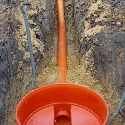 Pomarańczowy zbiornik z rurą w wykopie, widoczna łopata i fragment budynku w tle.