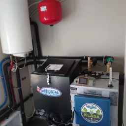 Instalacja kotła na paliwo stałe z zasobnikiem, widoczne rury, manometry, zbiornik wyrównawczy, sterownik i oznaczenia producenta Gregstal z informacją o 5 klasie i eco design