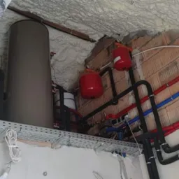 Instalacja zbiorników buforowych i rur grzewczych w budynku w stanie surowym, widoczne czerwone zbiorniki wyrównawcze, szare zasobniki, czarne rury izolowane oraz czerwone i niebieskie rury PEX...