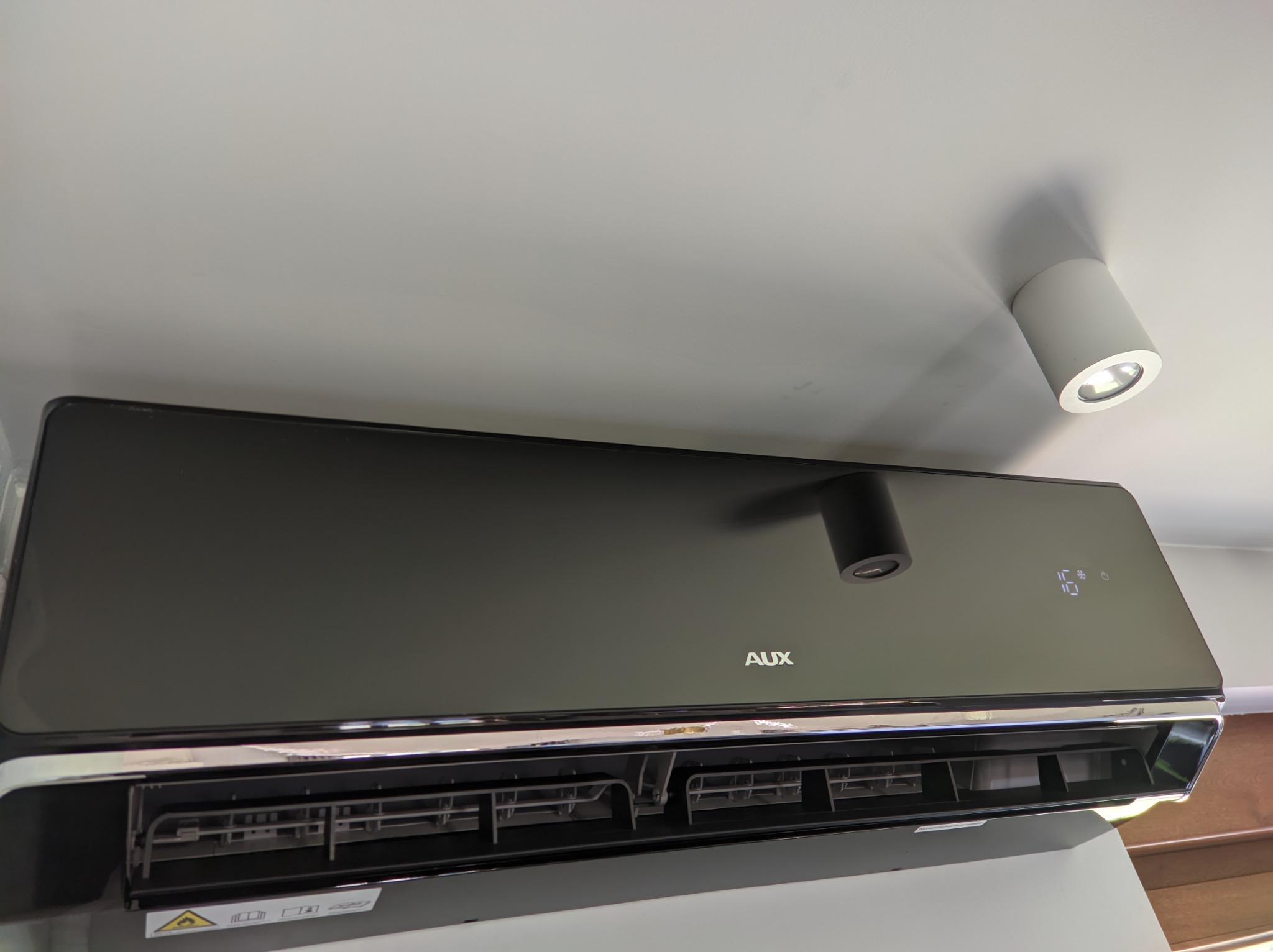 Czarny klimatyzator AUX zamontowany na ścianie, widoczny wyświetlacz temperatury i biała lampa sufitowa w tle.