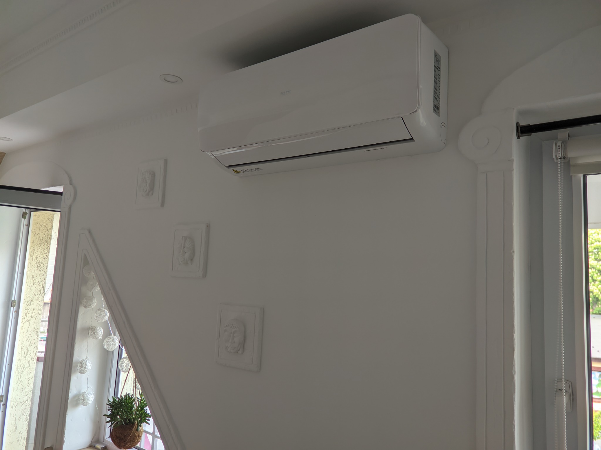 Biały klimatyzator marki AUX zamontowany na ścianie w jasnym pomieszczeniu z dekoracyjnymi sztukateriami i oknem z rośliną doniczkową.