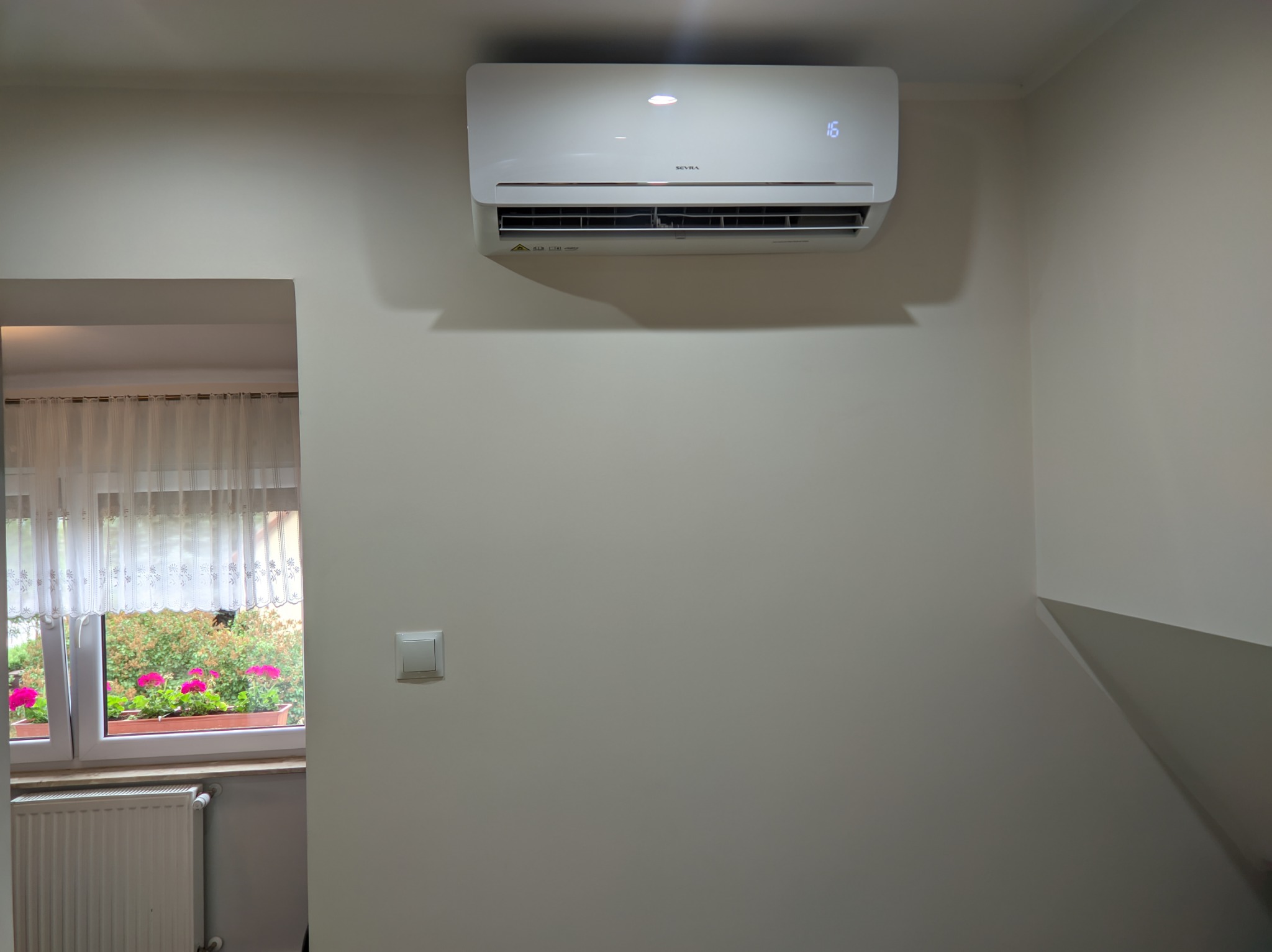Biała jednostka wewnętrzna klimatyzatora marki Seyra zamontowana na ścianie w pomieszczeniu z oknem, przez które widać kwiaty w doniczce.