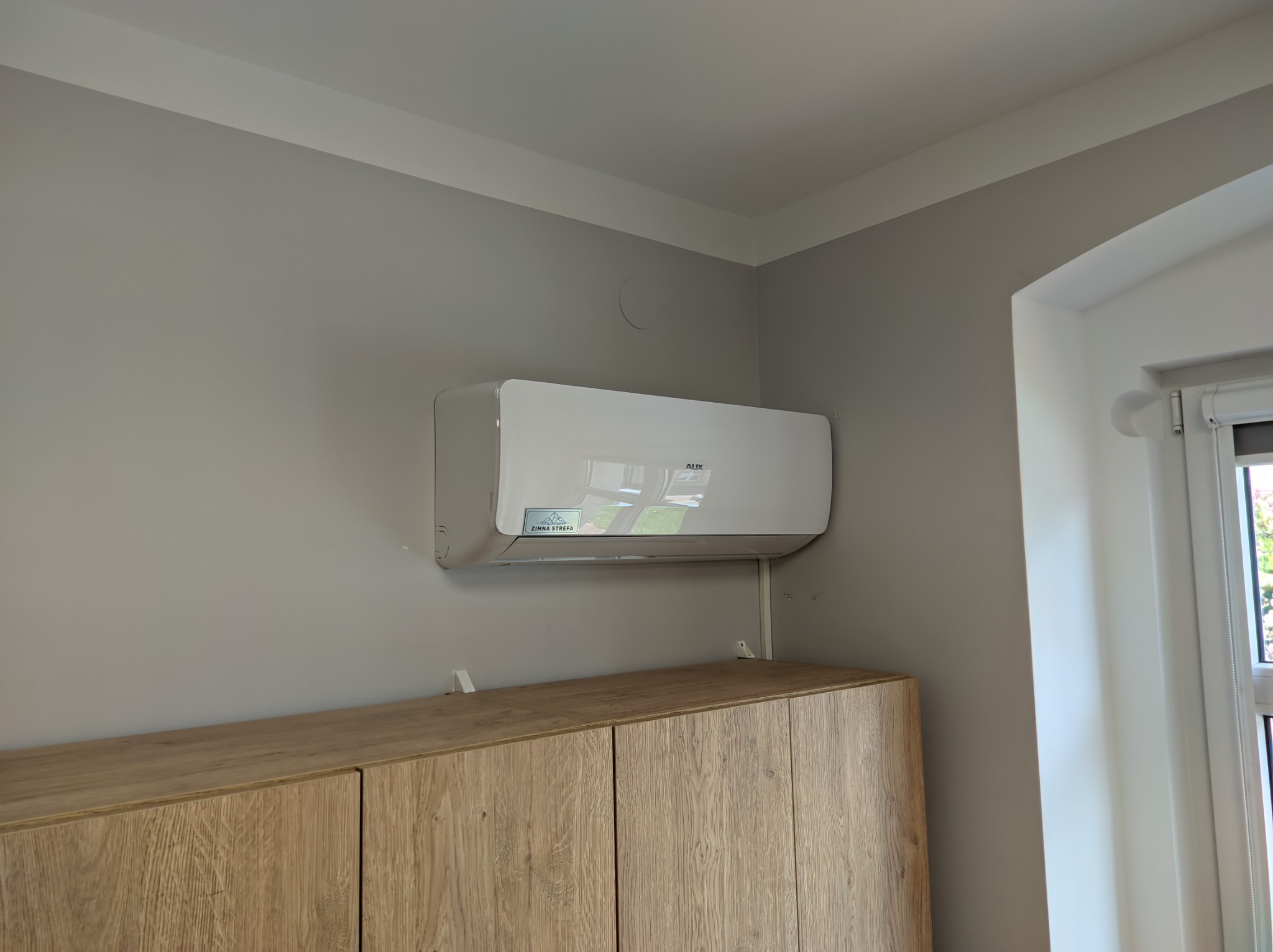 Biały klimatyzator AUX zamontowany na szarej ścianie nad szafką z jasnego drewna, widoczny fragment okna z roletą.