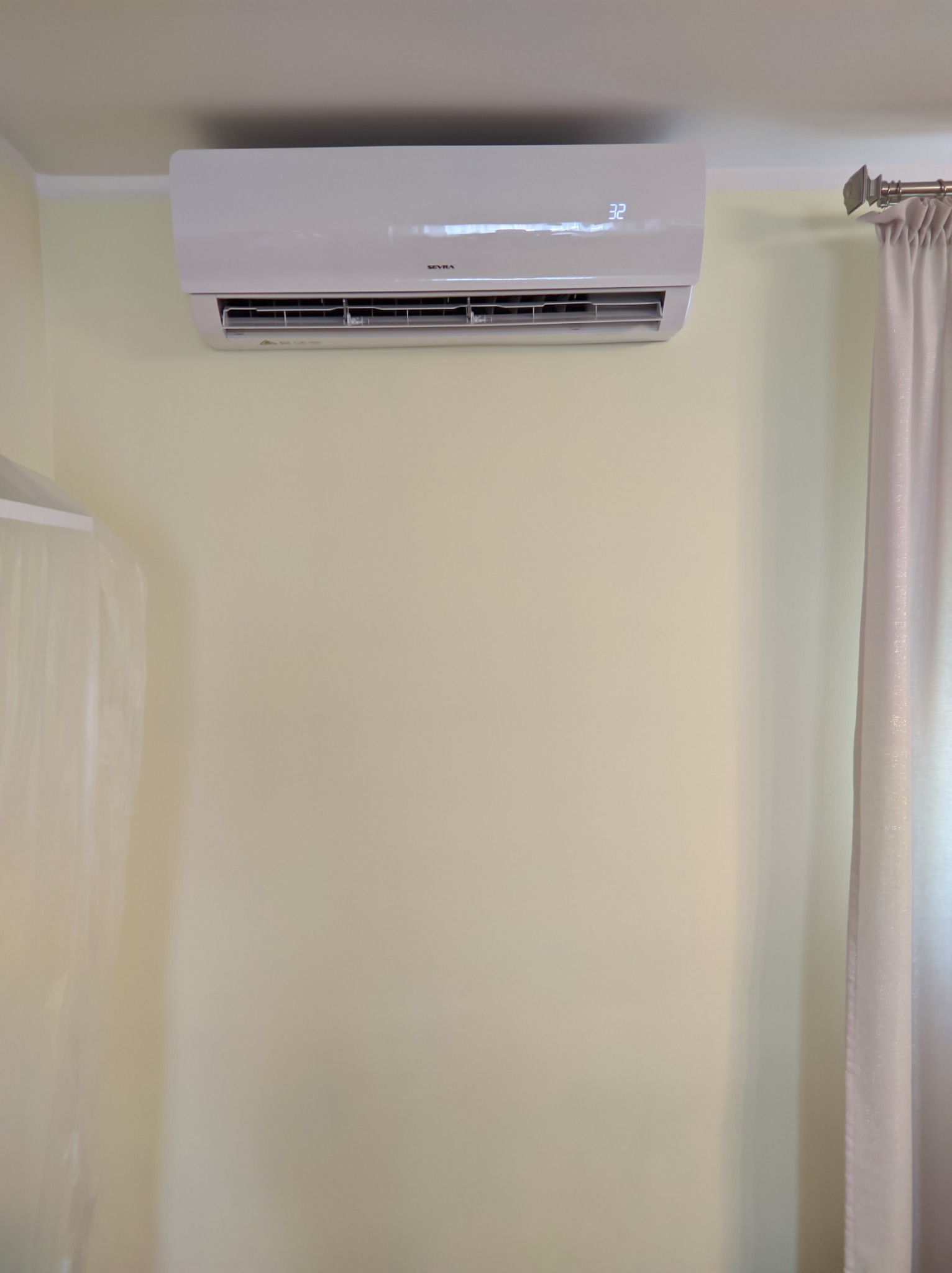 Biały klimatyzator ścienny z wyświetlaczem ustawionym na 32 stopnie, zamontowany na jasnożółtej ścianie, obok zasłony w kolorze ecru.