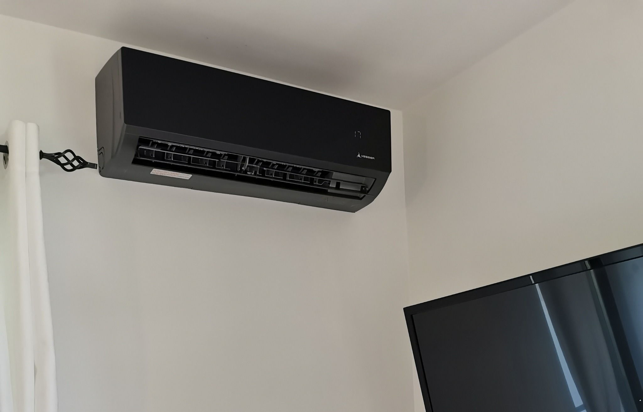 Czarny klimatyzator ścienny marki Vesser zamontowany w jasnym pomieszczeniu, widoczny fragment telewizora i zasłony.