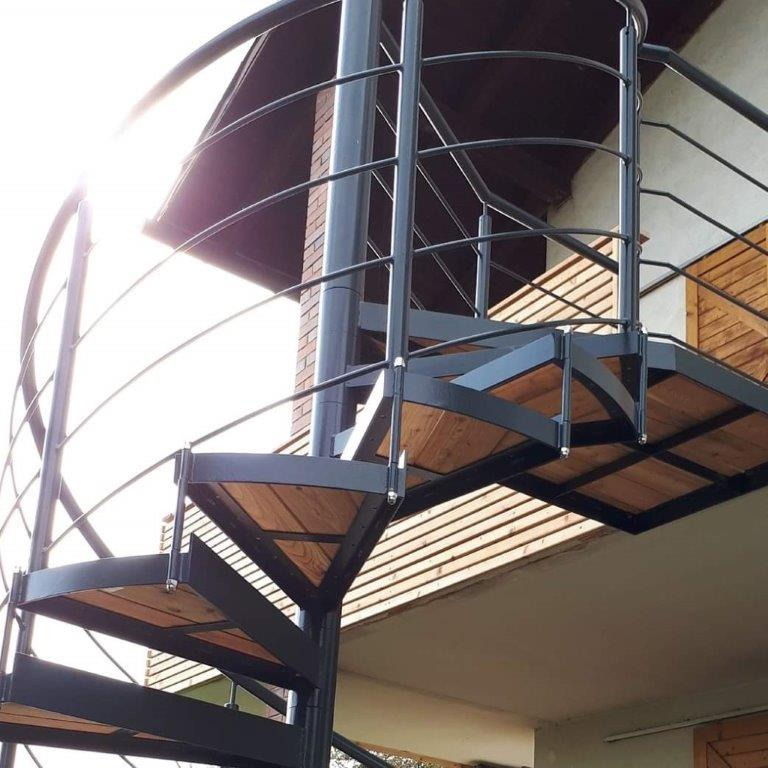 Zewnętrzne schody spiralne z czarnej stali z drewnianymi stopniami, prowadzące na balkon z drewnianą balustradą, widok z dołu na tle jasnego nieba.