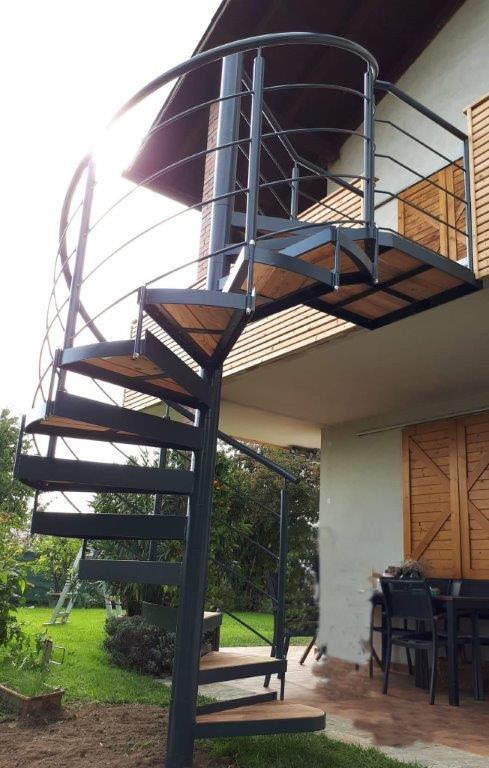 Zewnętrzne schody spiralne z czarną metalową konstrukcją i drewnianymi stopniami, prowadzące na balkon z metalową balustradą, na tle zielonego ogrodu i jasnej elewacji budynku.
