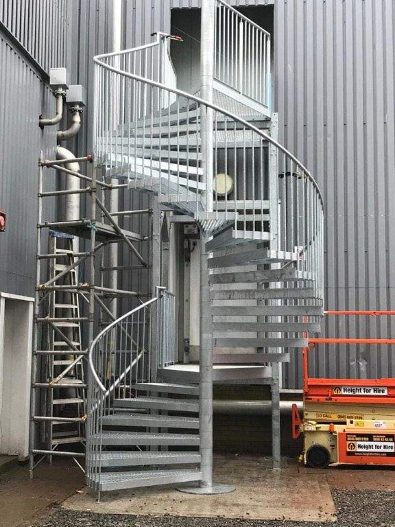 Zewnętrzne metalowe schody spiralne z zabezpieczeniami, prowadzące do drzwi na metalowej ścianie budynku przemysłowego, widoczne rusztowanie i podnośnik koszowy.