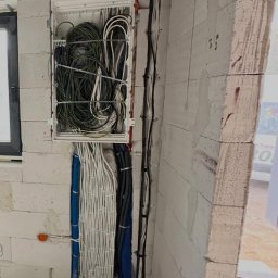 Wiktor Grabowski - Szczegółowe ujęcie rozbudowanej instalacji elektrycznej w surowym wnętrzu: otwarta rozdzielnica z gęsto ułożonymi przewodami, niebieskie peszle i kable biegnące po ścianach z bloczków oraz podłodze...