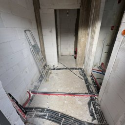 Wiktor Grabowski - Rozłożone czarne peszle z przewodami elektrycznymi i rury wodne na surowej podłodze w korytarzu budynku w trakcie budowy, z drabiną opartą o ścianę z białych bloczków.