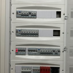 Wiktor Grabowski - Biała, otwarta rozdzielnica elektryczna z rzędem modułów zabezpieczających i wyłączników nadprądowych, oznaczonych jako 'REKUPERACJA', 'GARAŻ', 'STARY DOM', z czerwonym wskaźnikiem.