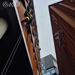 ASAP Maciej Książczyk - Pracownicy alpinistyczni na linach podczas prac na wysokościach na elewacji budynku o rdzawym kolorze, widok z dołu.
