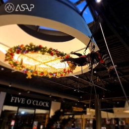 ASAP Maciej Książczyk - Pracownik w kasku, zabezpieczony linami, montuje świąteczną dekorację z bombkami i lampkami pod sufitem galerii handlowej, widoczne sklepy Five O'Clock i Pieca Rodem.