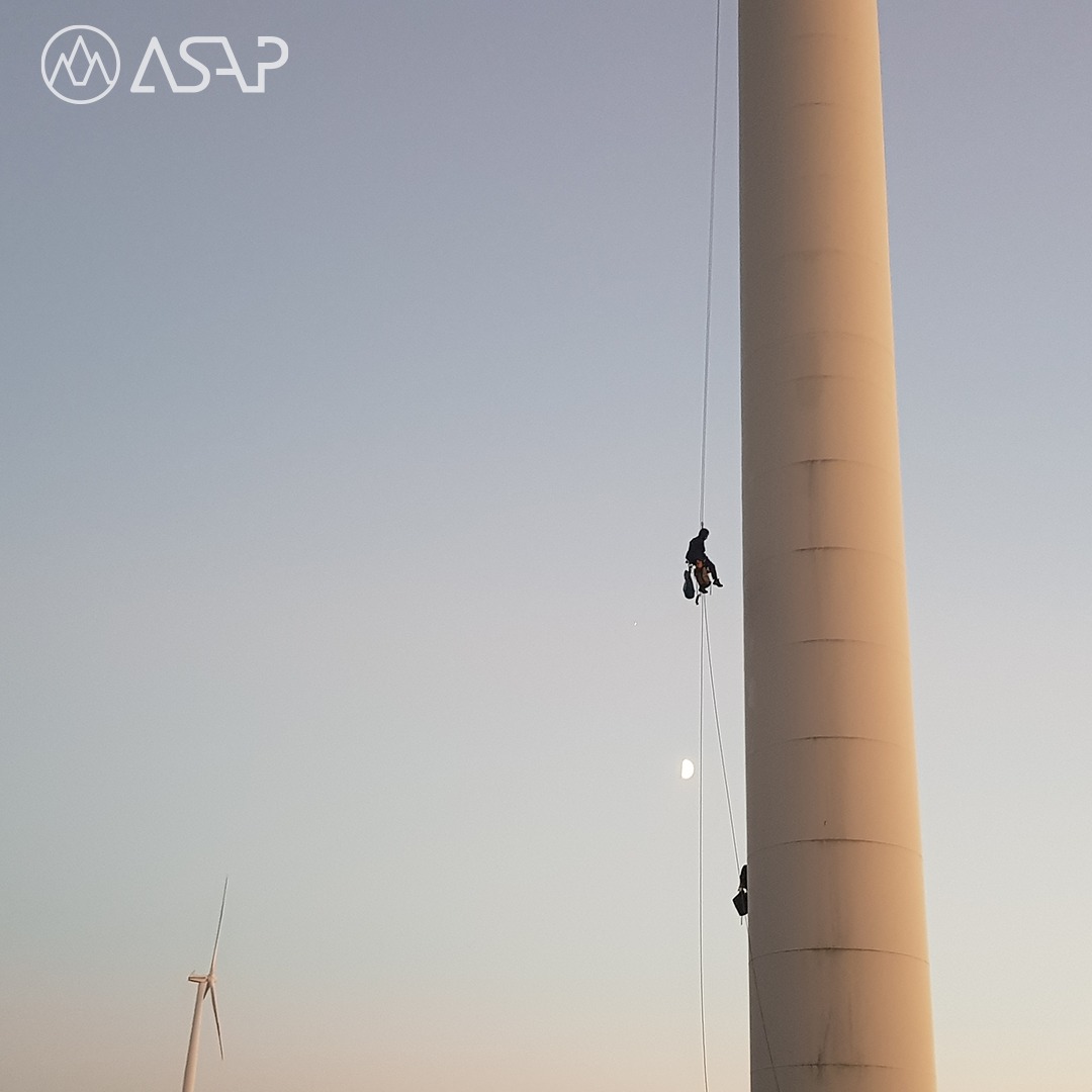Dwóch pracowników na linach podczas pracy na turbinie wiatrowej; w tle widoczna inna turbina i jasne niebo.