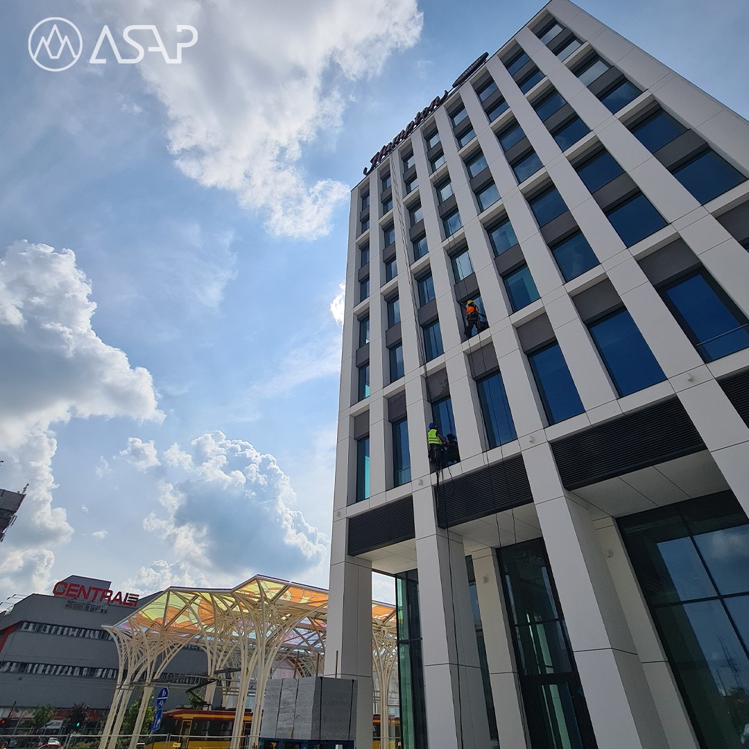 Dwóch alpinistów na linach zabezpieczających, pracujących na wysokościach przy elewacji nowoczesnego, wysokiego budynku biurowego z licznymi oknami, widok od dołu w słoneczny dzień, w tle błękitne...