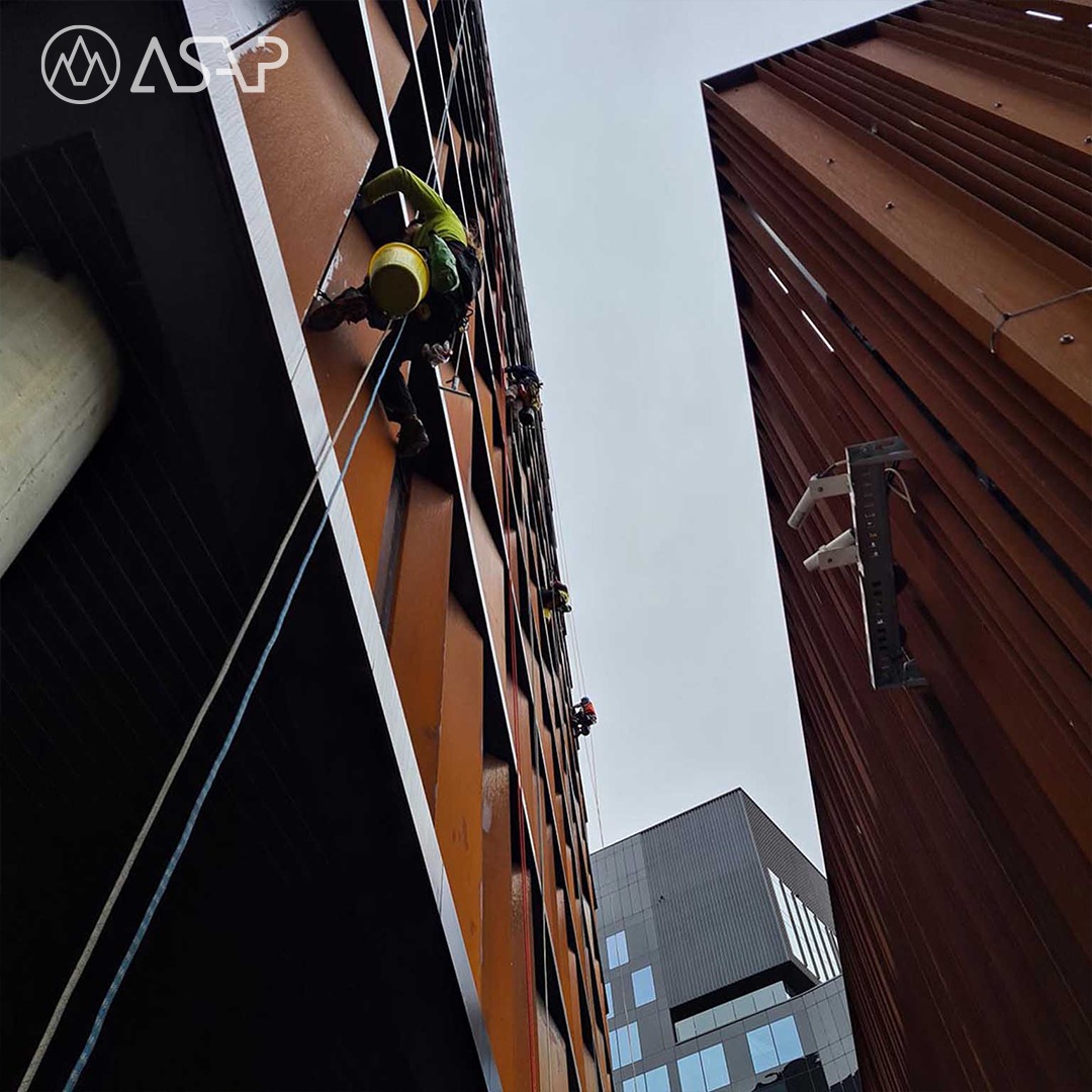 Pracownicy alpinistyczni na linach podczas prac na wysokościach na elewacji budynku o rdzawym kolorze, widok z dołu.