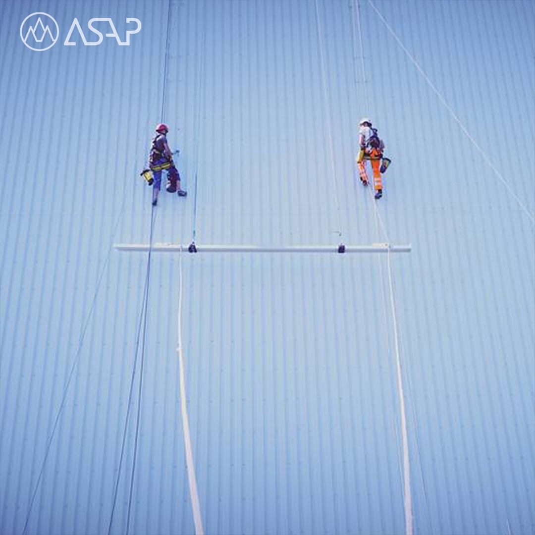 Dwóch pracowników na linach zabezpieczających, wykonujących prace na wysokości na dużej, błękitnej, blaszanej ścianie budynku z widocznym logo ASAP w lewym górnym rogu.