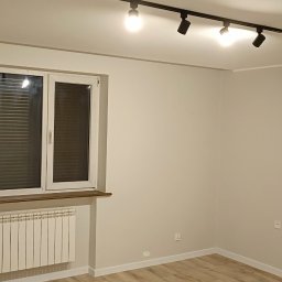 Usługi remontowo budowlane Hubert Sznajder - Pomalowane na kremowo wnętrze pokoju z drewnianą podłogą, białym grzejnikiem pod oknem z żaluzjami, oraz czarną lampą sufitową z czterema reflektorami.