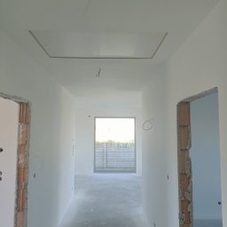 Usługi remontowo budowlane Hubert Sznajder - Korytarz w nowym domu w trakcie wykańczania, ściany świeżo pomalowane na biało, widoczne surowe ościeżnice drzwiowe i otwór w suficie.