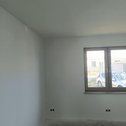 Usługi remontowo budowlane Hubert Sznajder - Wnętrze pokoju po świeżym pomalowaniu ścian na biało, z widocznym oknem w drewnianej ramie i przygotowanym podłożem pod panele.