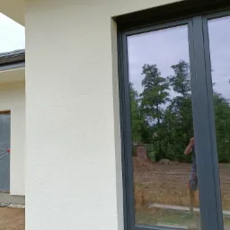 Nowe, grafitowe okno PCV zamontowane w elewacji budynku, widoczne odbicie drzew i osoby robiącej zdjęcie.