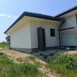 Elewacja budynku po nałożeniu tynku, widoczne rusztowanie, okno i tymczasowe zabezpieczenie wejścia.