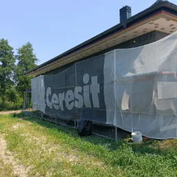 Dom w trakcie ocieplania, widoczne rusztowania, siatka ochronna z logo Ceresti, czarne płyty izolacyjne na ścianie, fragment ocieplonego poddasza z widocznymi kołkami.