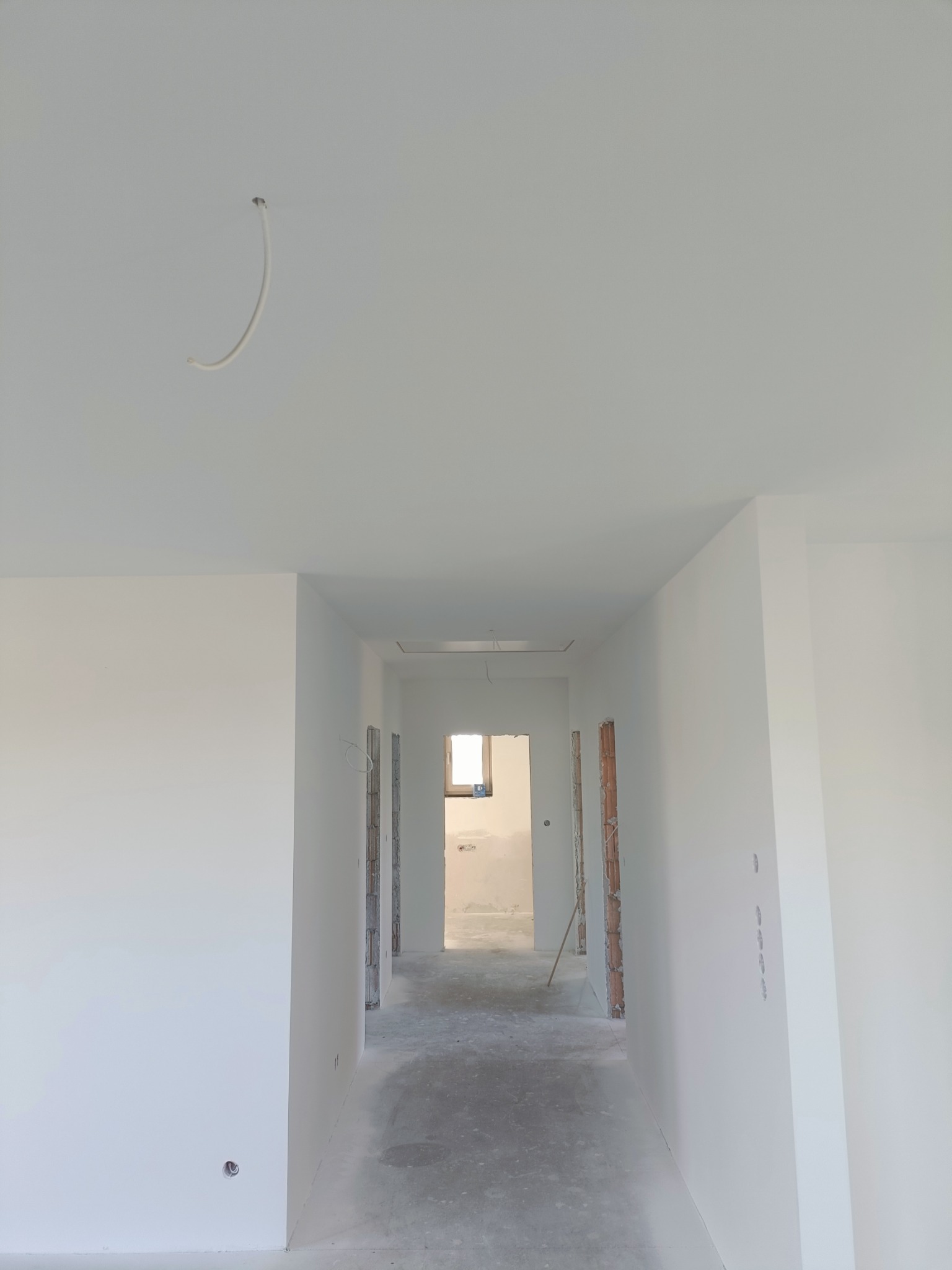 Wnętrze w trakcie wykańczania: białe ściany po gładzi, surowy beton na podłodze, widoczne kable elektryczne wystające z sufitu i ściany, oraz fragmenty muru w otworach drzwiowych.