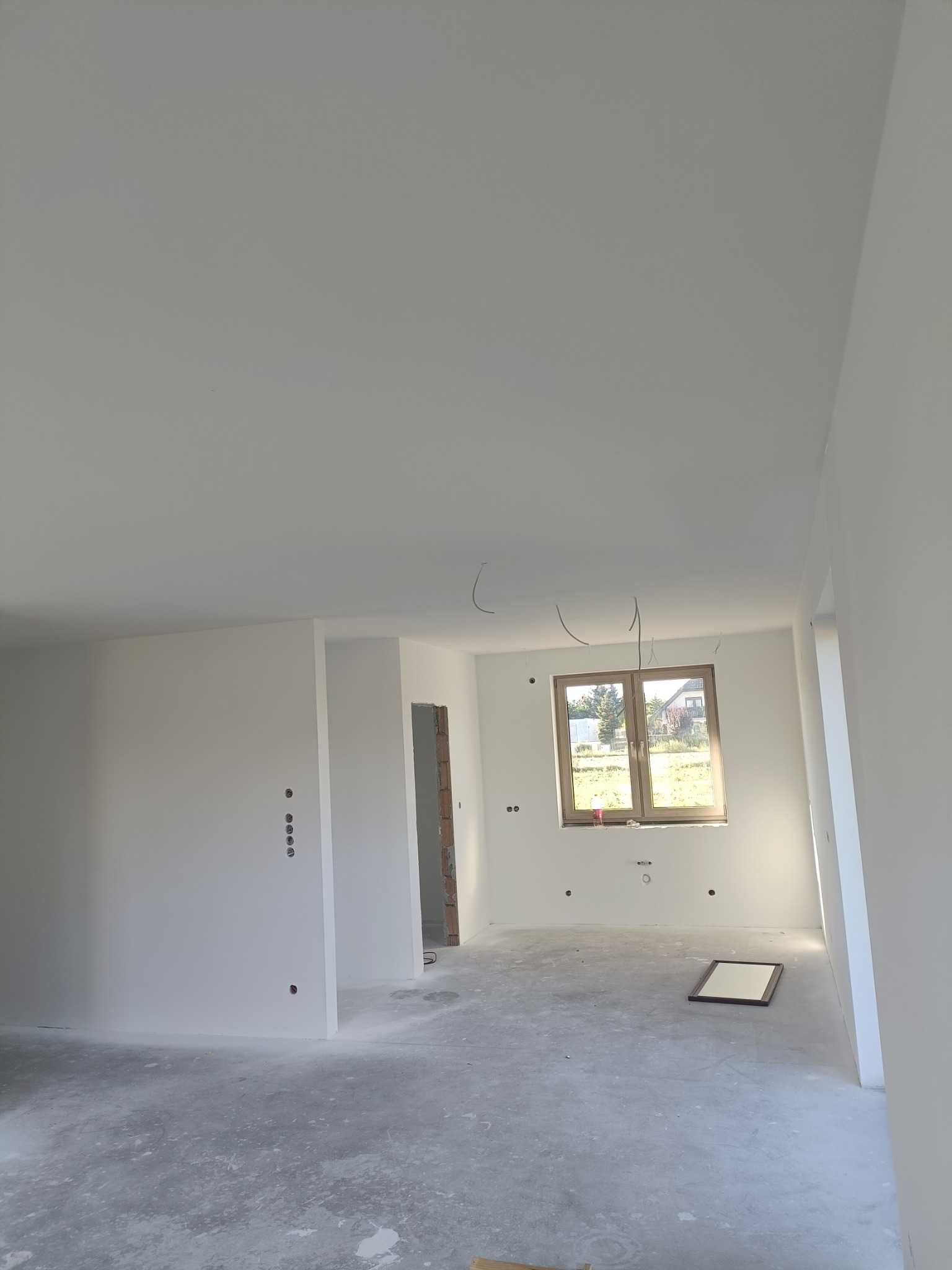 Wnętrze po gładziowaniu i malowaniu na biało, widoczne wystające przewody elektryczne, surowa podłoga, okno z widokiem na zewnątrz, fragment ramy obrazu na podłodze.