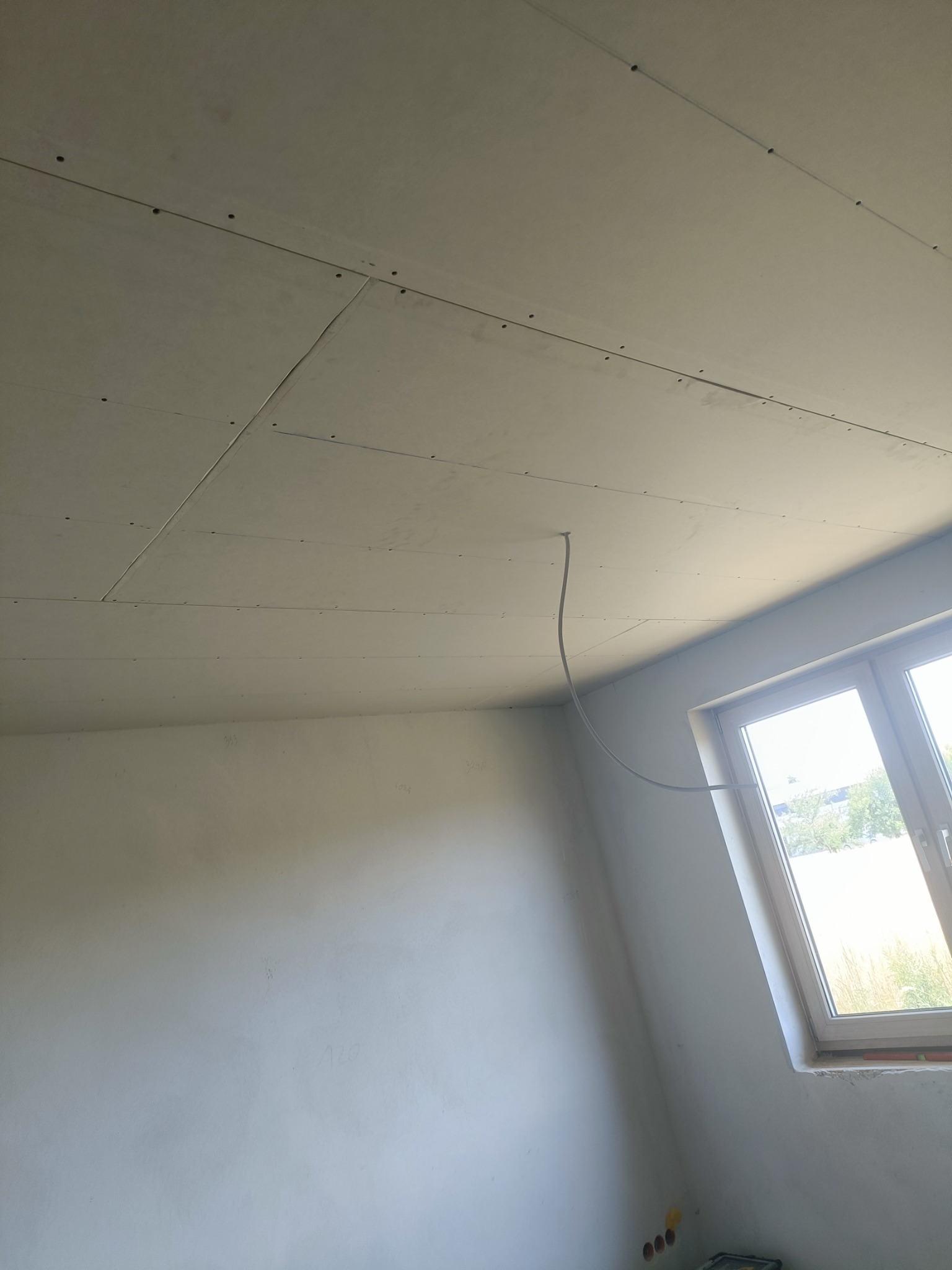Płyty kartonowo-gipsowe zamontowane na suficie w pomieszczeniu w trakcie remontu, widoczne łączenia płyt i przewód elektryczny wystający z sufitu obok okna.