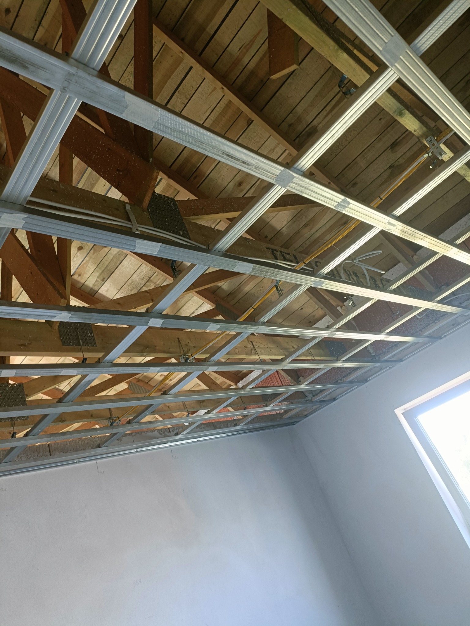 Stelaż z profili metalowych pod zabudowę sufitu z płyt gipsowo-kartonowych, widoczne drewniane elementy konstrukcyjne dachu i instalacje, fragment ściany z oknem w tle.
