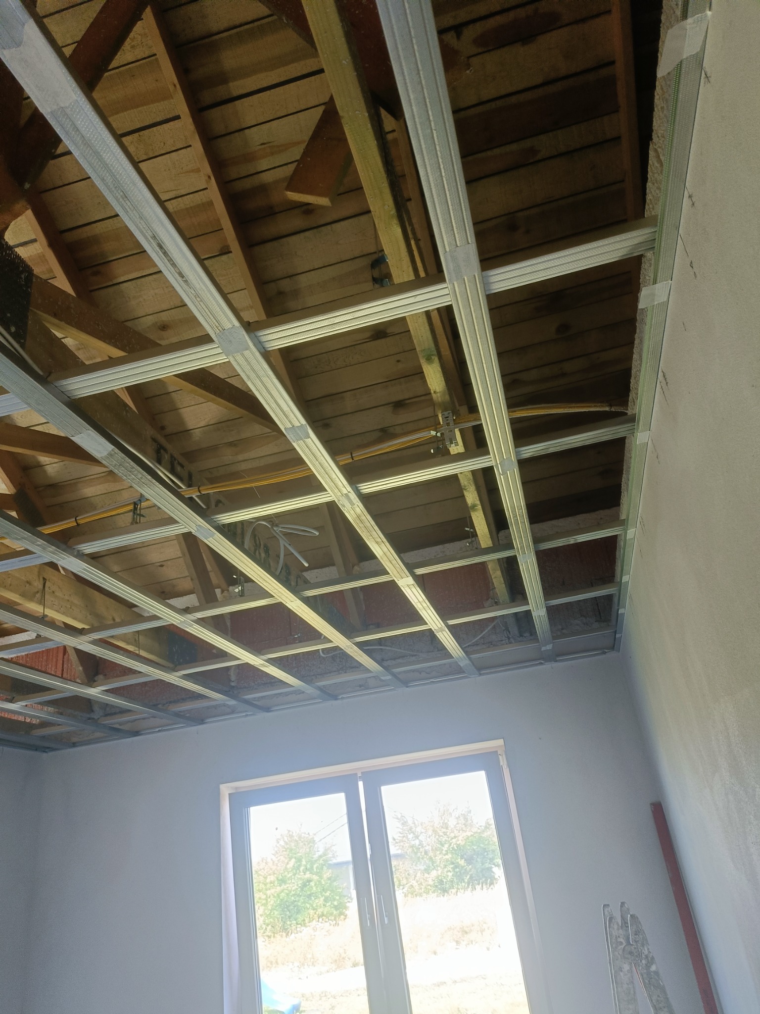 Metalowa konstrukcja podwieszanego sufitu z płyt kartonowo-gipsowych zamontowana w pomieszczeniu z oknem, widoczne drewniane belki stropowe i instalacja elektryczna.