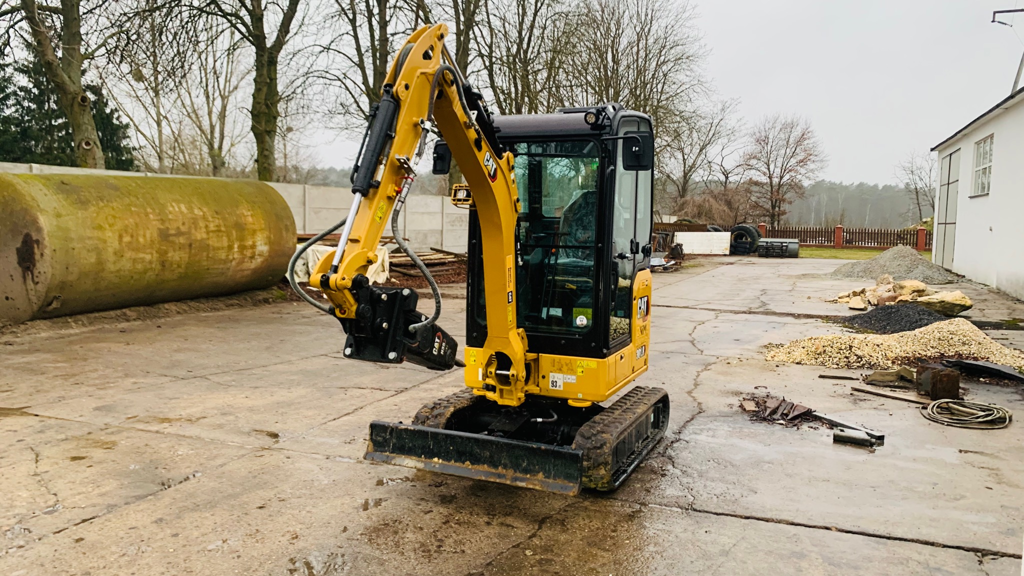 Żółta mini koparka Caterpillar z hydraulicznym młotem na betonowym placu budowy, widoczny duży betonowy cylinder i sterta gruzu w tle.