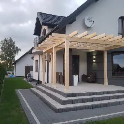 Nowoczesny dom z drewnianą pergolą nad tarasem, widoczne schody z kostki brukowej, trawnik i tuje w tle.