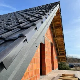 Nowy dach z czarnej dachówki ceramicznej, widok z dołu na fragment konstrukcji z cegły i drewnianą więźbę, w tle błękitne niebo.