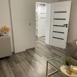 Usługi Remontowo Budowlane Sebastian Piasecki - Nowoczesne wnętrze mieszkania z podłogą imitującą drewno, białymi drzwiami z poziomymi przeszkleniami i szklanym stolikiem kawowym z dekoracjami.