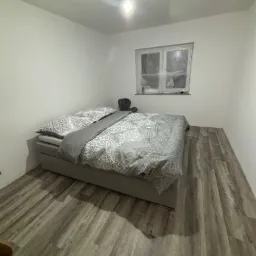 Pokój z nową podłogą z paneli w odcieniach szarości, łóżko z szarą pościelą, okno zakryte folią malarską, surowa żarówka na suficie.