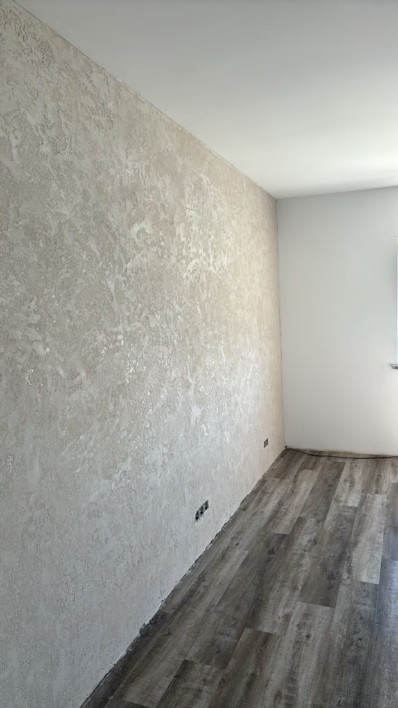 Ściana z dekoracyjnym tynkiem w odcieniach szarości i beżu, panele podłogowe imitujące drewno, widoczne gniazdka elektryczne w ścianie, narożnik pokoju z białymi ścianami.