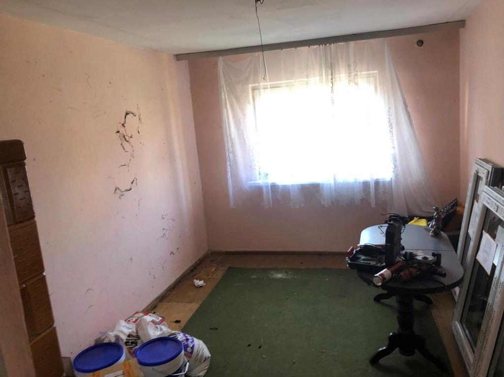 Pokój w trakcie przygotowań do remontu: ściany w odcieniach różu z widocznymi śladami po wcześniejszych dekoracjach, okno zasłonięte prześwitującą firanką, na podłodze zielony dywan i worki...