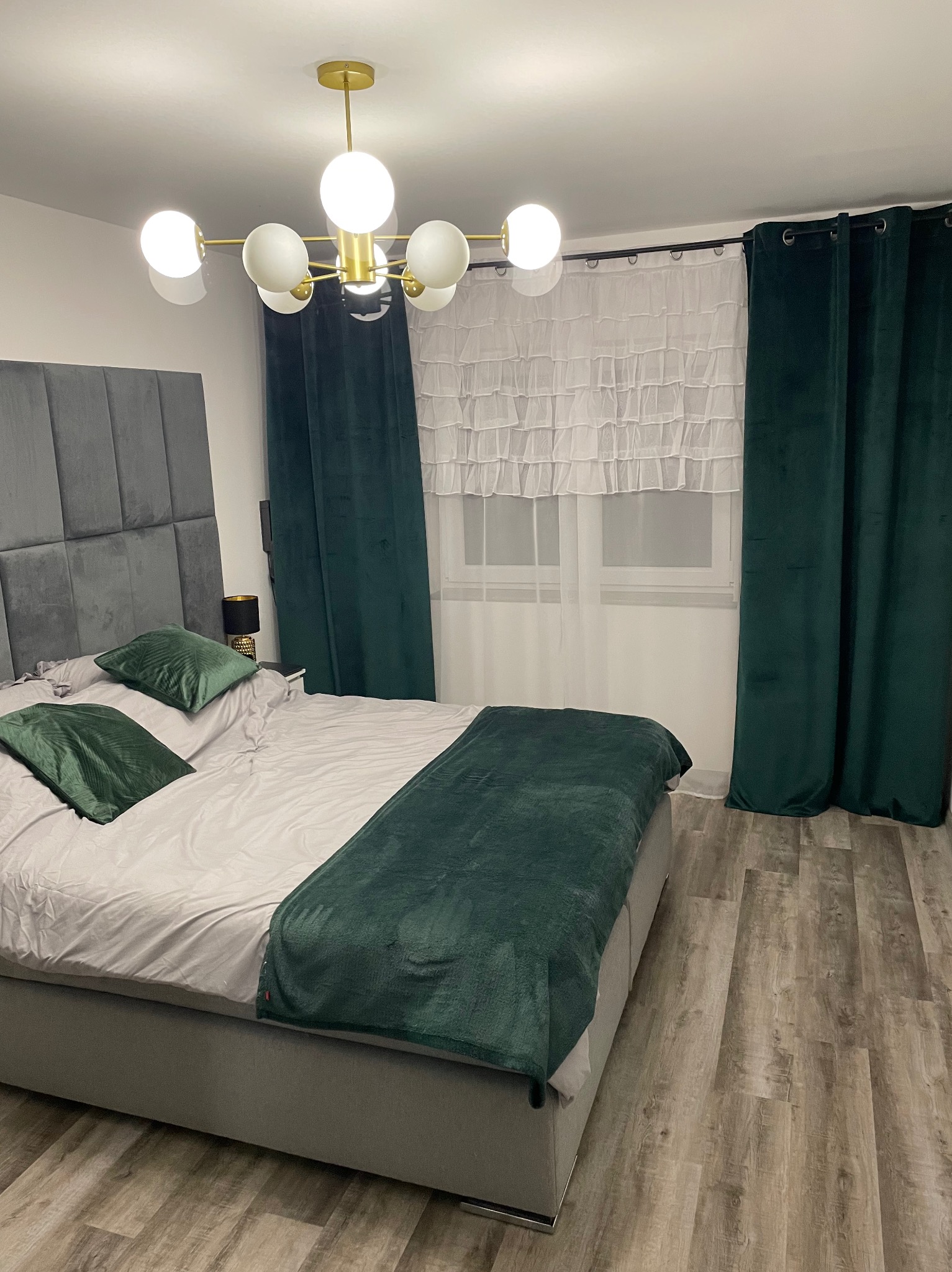 Sypialnia z szarym tapicerowanym łóżkiem, zielonymi poduszkami i narzutą, oraz ciemnozielonymi zasłonami po bokach okna z białą firanką, oświetlona nowoczesną lampą sufitową z białymi kulami.