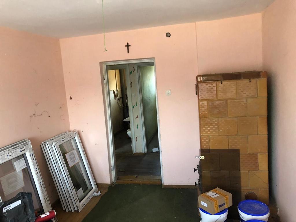 Wnętrze pokoju z przygotowanymi do montażu oknami, widoczny piec kaflowy i wejście do łazienki, ściany w odcieniu brudnego różu.