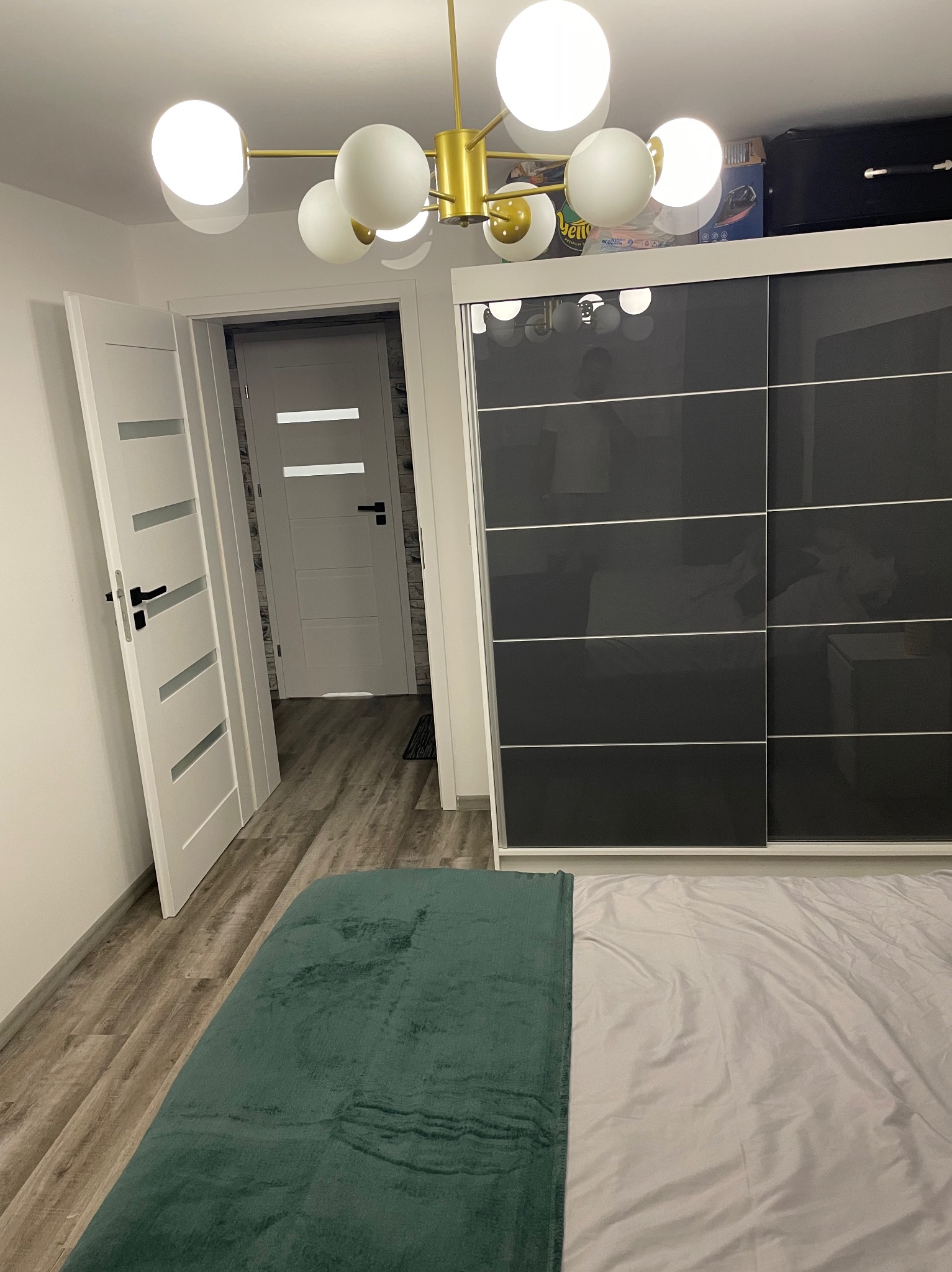 Nowoczesna sypialnia z przesuwną szafą w kolorze grafitowym, widoczna podłoga z paneli imitujących drewno, białe drzwi z poziomymi przeszkleniami i designerska lampa sufitowa z kulistymi kloszami.