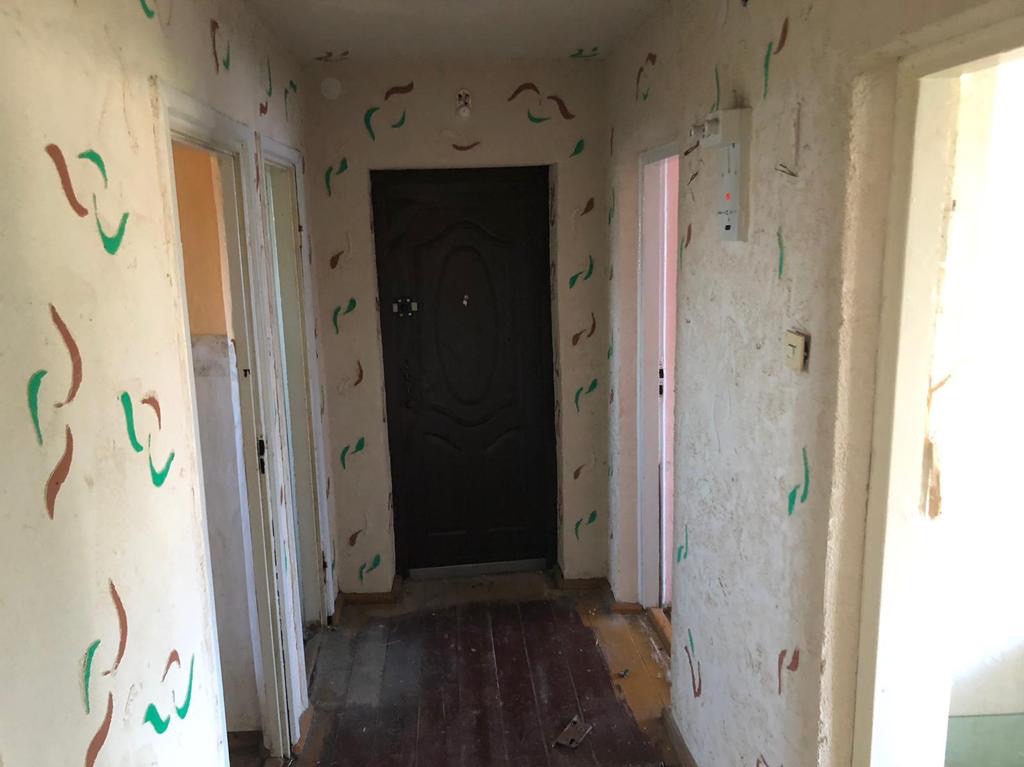 Korytarz przedpokoju z ciemnymi drzwiami wejściowymi, ściany pokryte wzorem w kształcie przecinków w kolorze brązowym i zielonym, widoczne ślady użytkowania i zniszczenia.